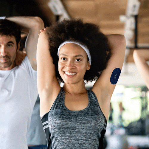 A black woman wearning a grey tank top extending her arms over her head with her elbows bent. She's wearing a CGM and a Signos sports cover on her left arm.