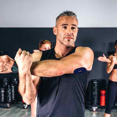 A middle-aged white man wearing a black tank top with a CGM and a Signos sports cover on his left tricep. He's taking a group exercise class and stretching his left deltoid by pulling his left arm across his chest and pulling on his left forearm with his right hand.