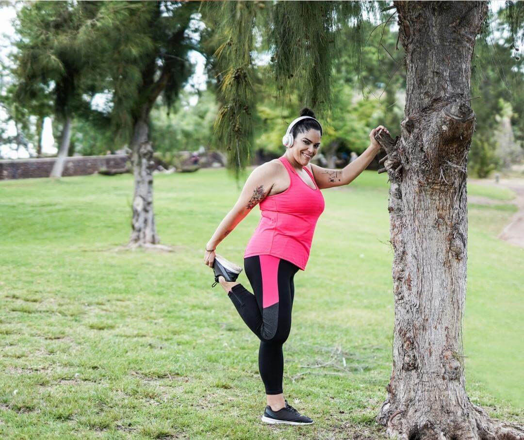 a heavyset woman wearing wireless headphones and stretching her quadriceps while holding onto a tree for balance