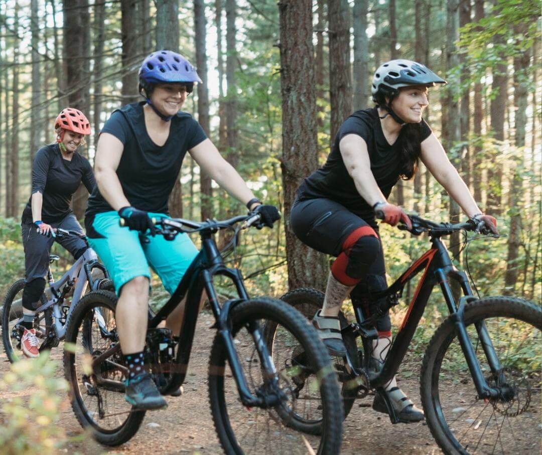 three women riding bikes on a forest trail