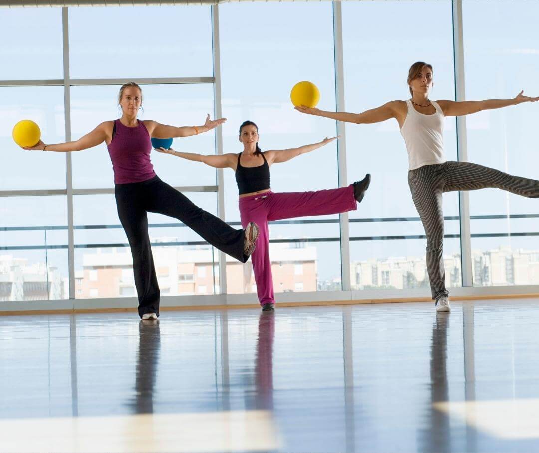 three women in an exercise class standing on one foot with both arms outstretched, a ball in one hand