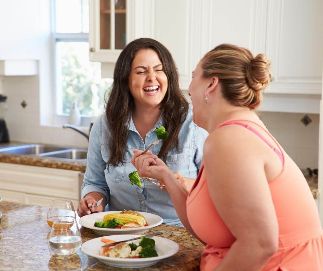 two heavyset women eating vegetables in a kitchen