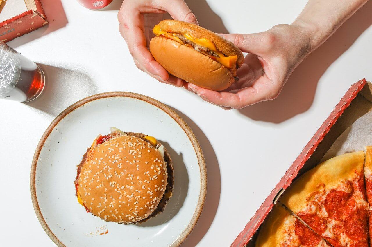 person-holding-cheeseburger-with-pizza-and-soda-on-the-side