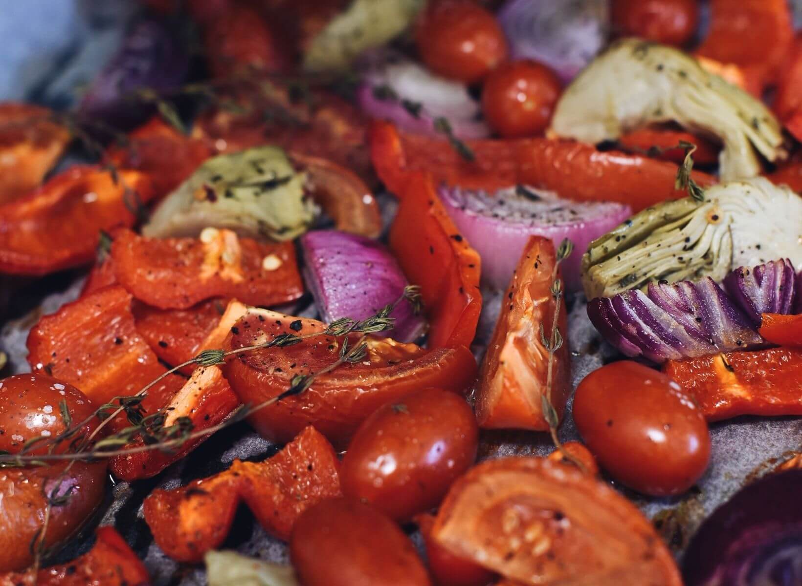 Close up shot of cherry tomatoes, sliced tomatoes, red onions, artichoke hearts, and thyme sprigs on a sheet pan from the Mediterranean chicken sheet pan dinner recipe