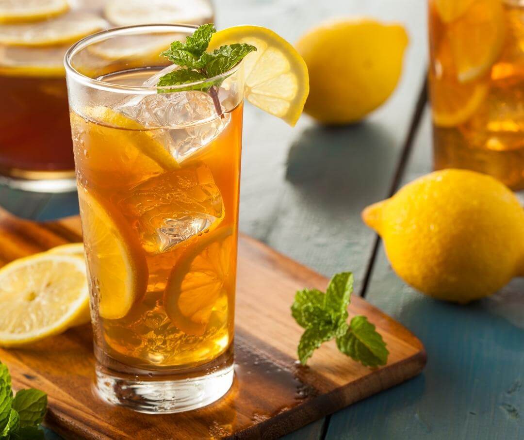 glass of iced tea with a lemon slice and sprig of mint, with whole lemons in the background