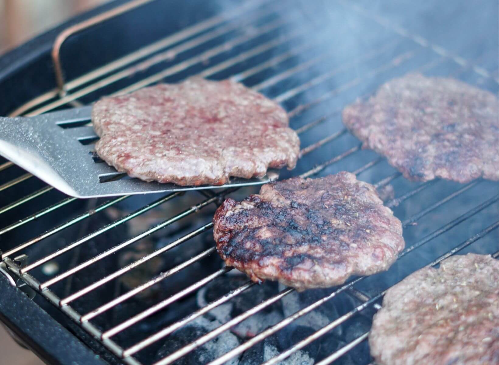 Close up shot of a burger on a spatula. Other burger patties are cooking on the grill grates.