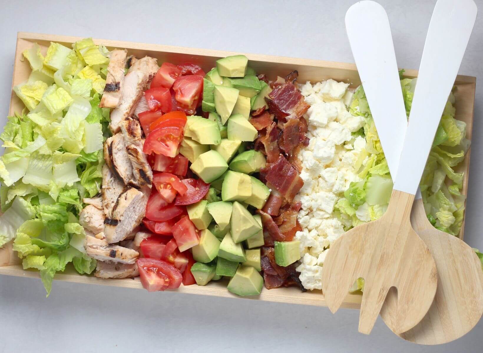 Cobb salad on a wooden serving dish with salad serving spoons on the side