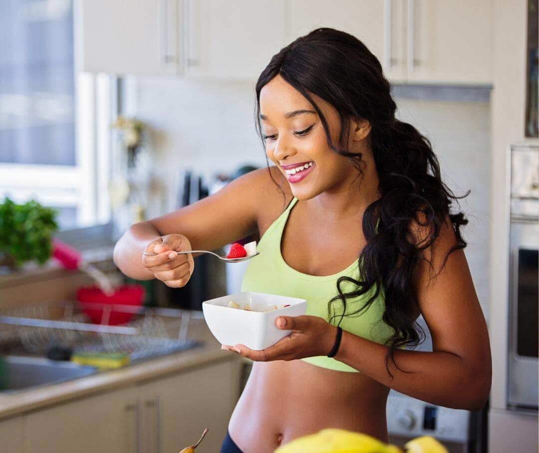 woman standing in a kitchen eating a bowl of berries and yogurt